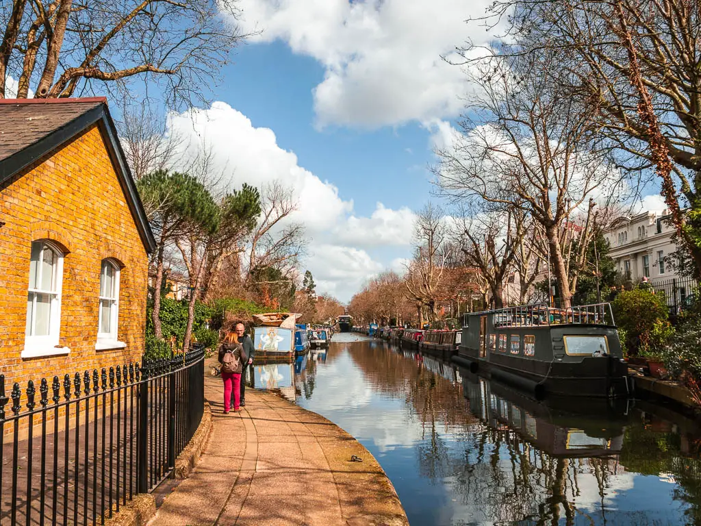 The regents canal running straight ahead, with the path on the left, at the start of the walk. There is a small brick bundling on the left of the path with a black metal railing in front. There are a couple of people standing on the path. 