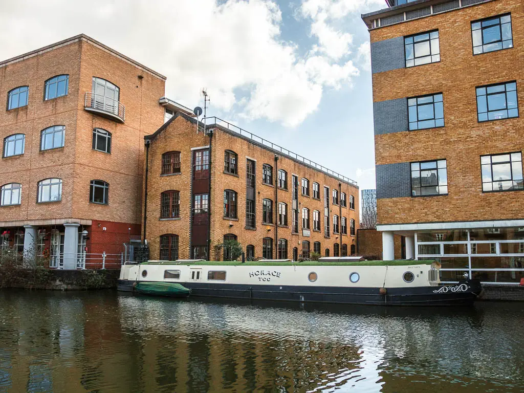 Looking across the canal to large brown bricked buildings and a moored white and green barge.