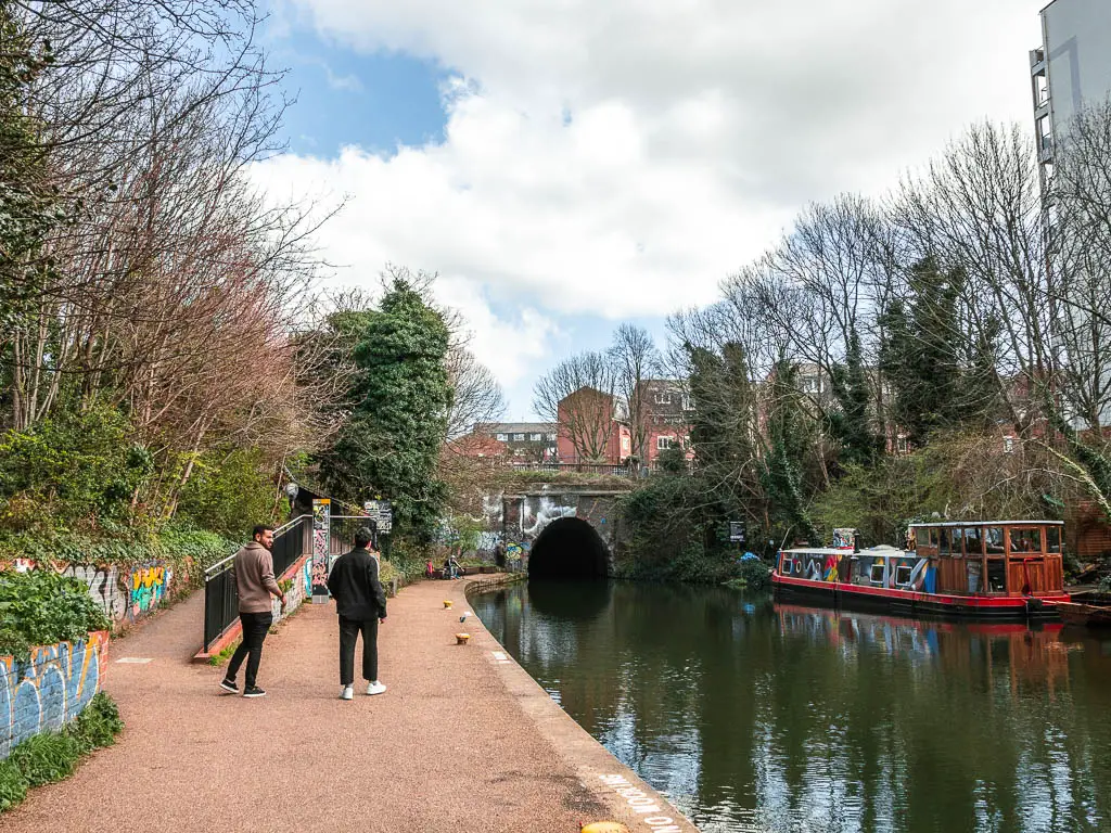 The path with the canal to the right and an archway bridge up ahead where the path stops. There are two people walking on the path and a sloped walking leading off it to the left. 