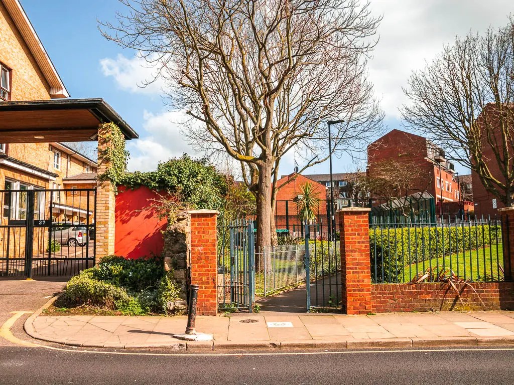 Looking across the road to brick and metal gates, leading onto a pathway next to a small green.