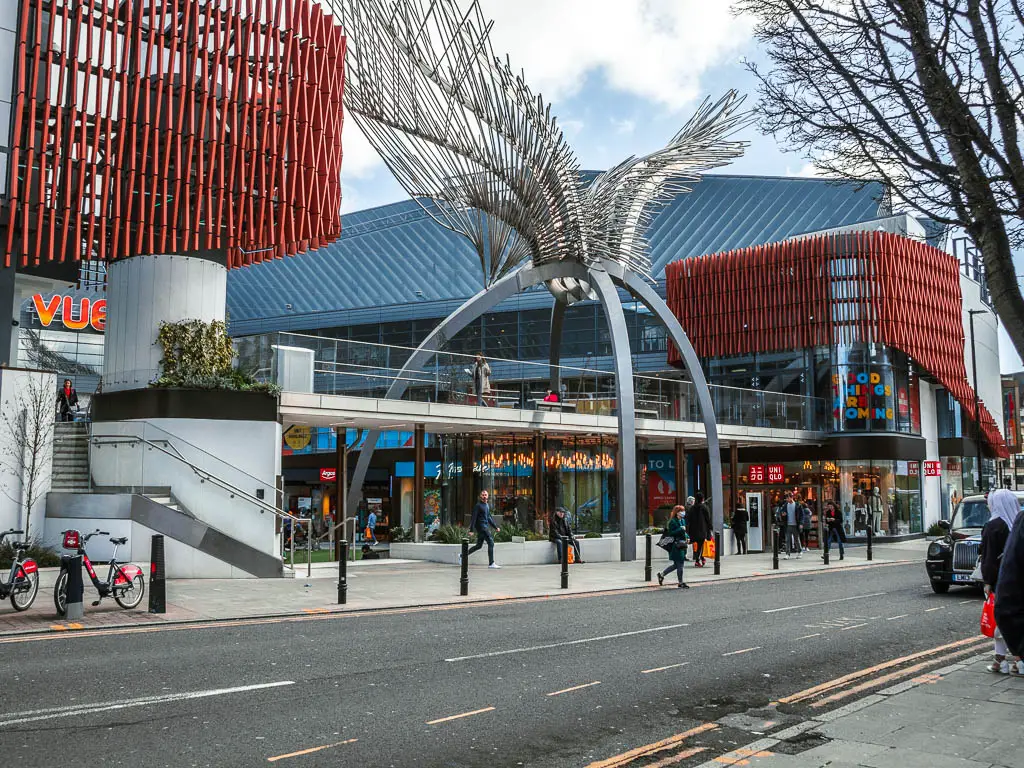 A large shopping centre across the road, with lots of people walking about.