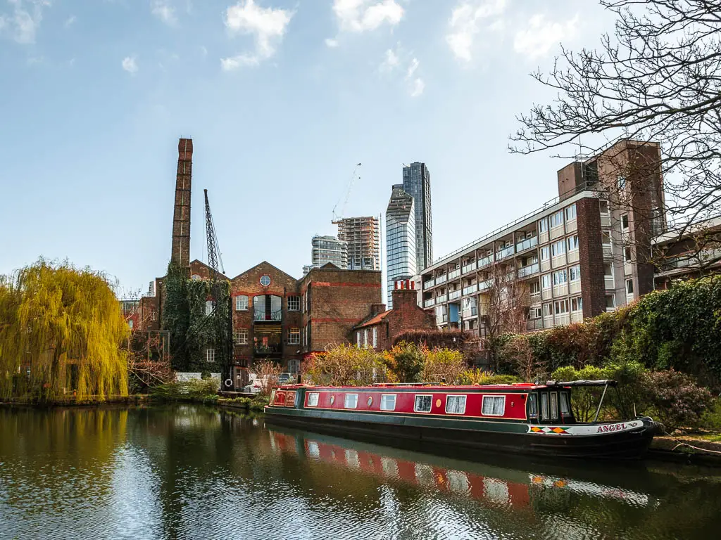 Looking across the water to an old factory warehouse, and apartment blocks on the walk along the Regents Canal in London. there is a red barge moored to the side, and skyscrapers in the distance.