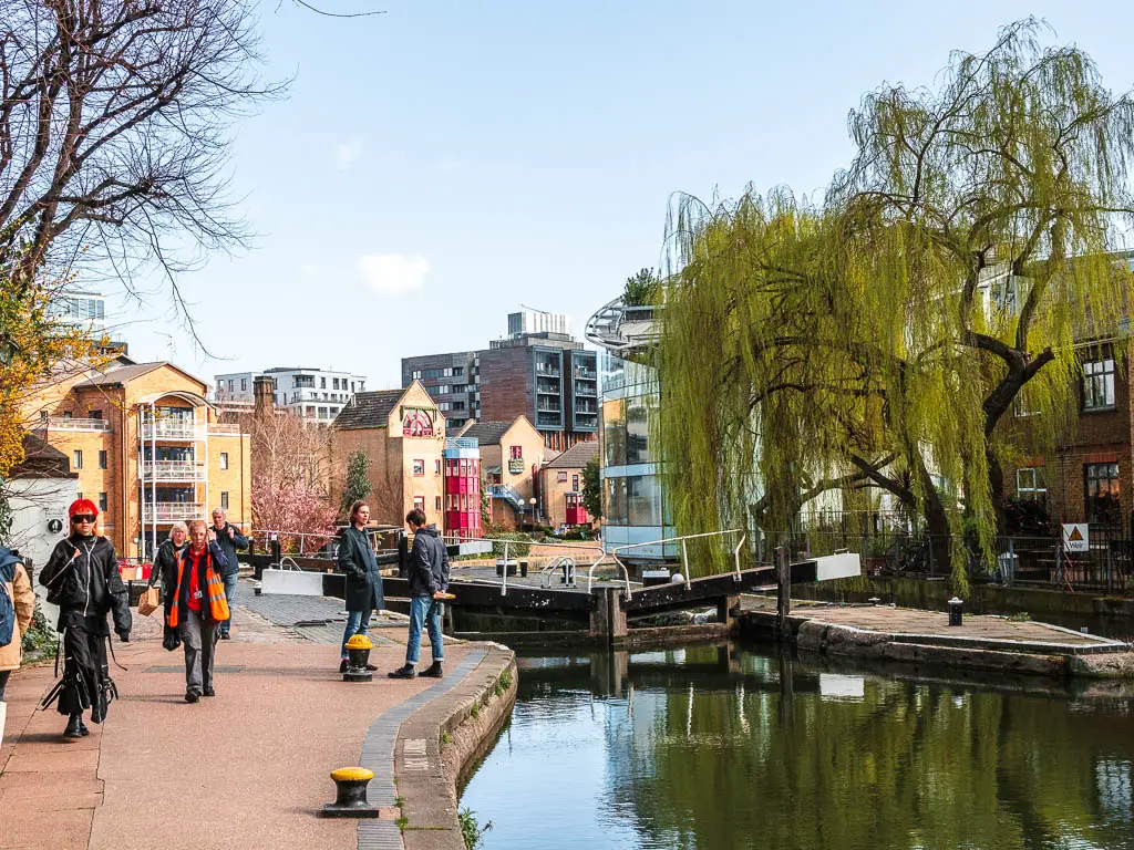 The Regents canal towpath with lots of people walking on it. There is a lock in the canal, and apartment buildings up ahead.