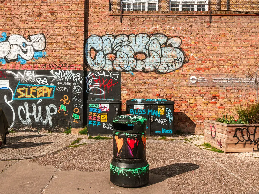 A black bin with graffiti, in front of a brick wall with graffiti. 