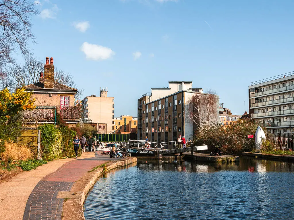 The path on the left of the water, with a lock ahead, and an assortment of apartment buildings.