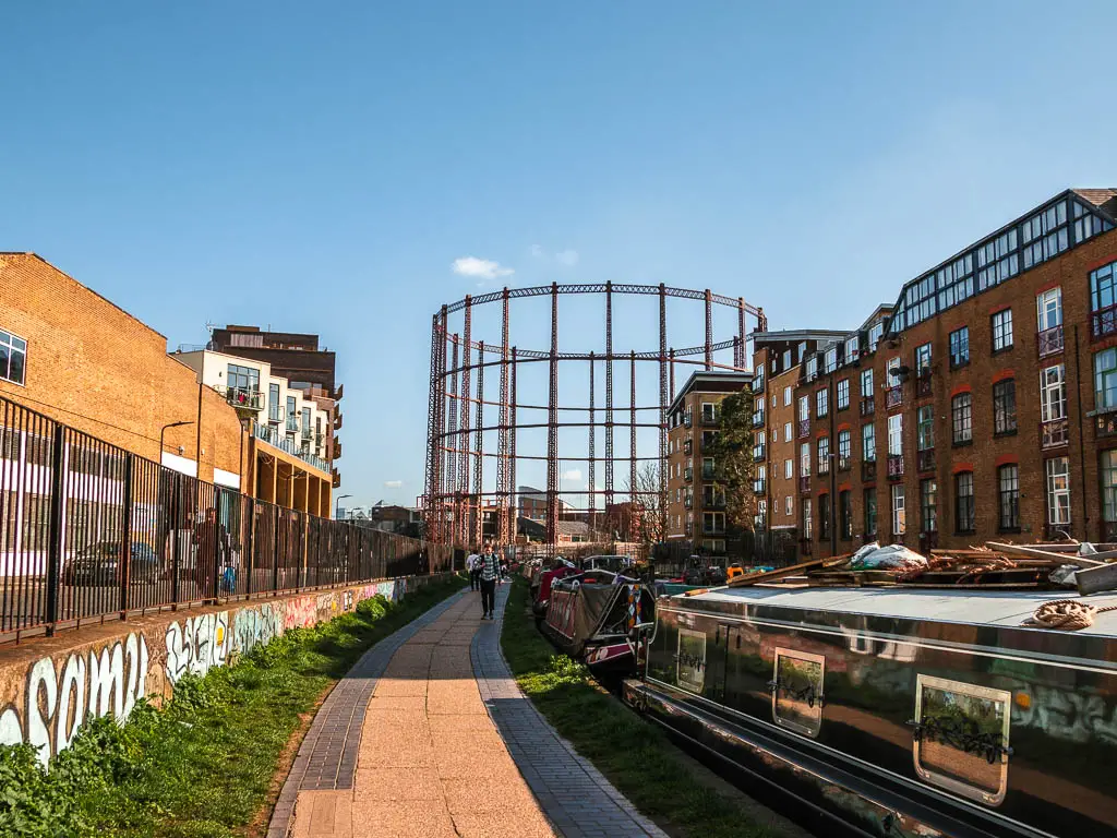 The path with the road to the left on the other side of black railings, and barges moored to the side on the right. There is a large metal gas holder straight ahead.