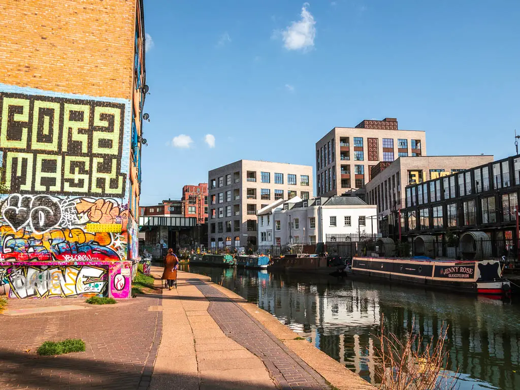 Walking along the oath next to the Regents Canal in London, with an assortment of buildings on the other side, and a wall with graffiti on this side. 