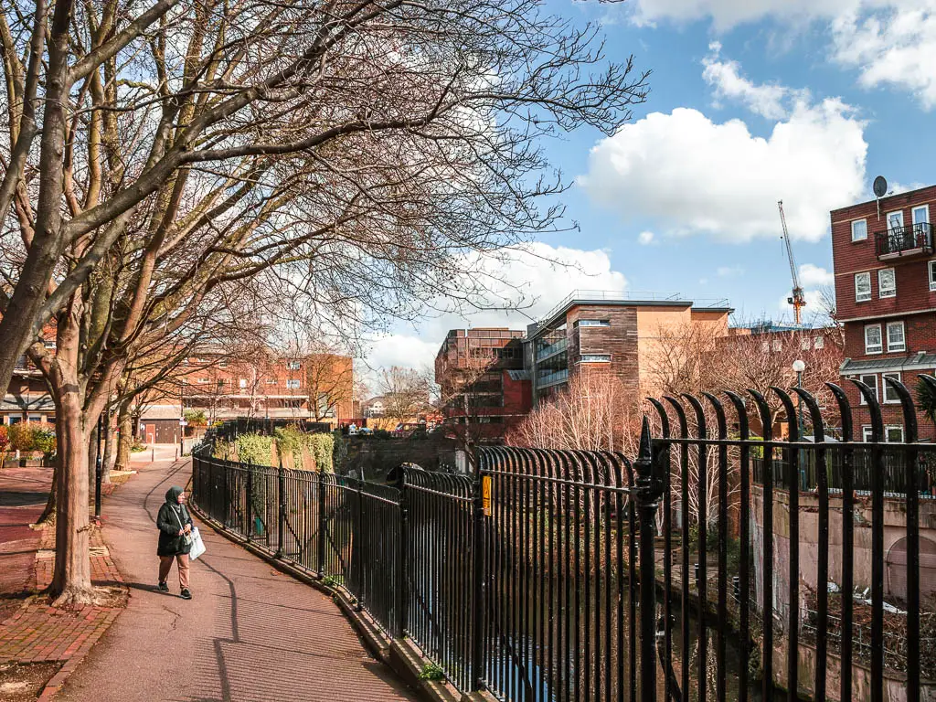 A path on the left, with black metal railings to the right, and the canal on the other side, down down. There is a person walking on the path.