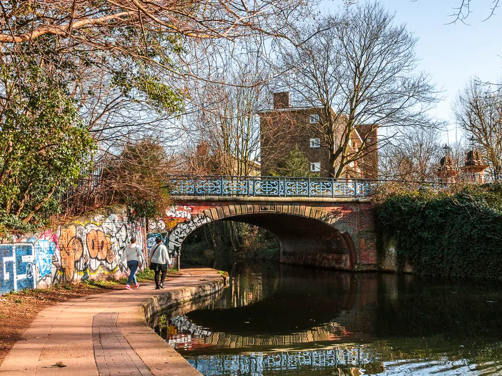 A path leading under a bridge next to the regents canal, with a couple of people walking on the path.