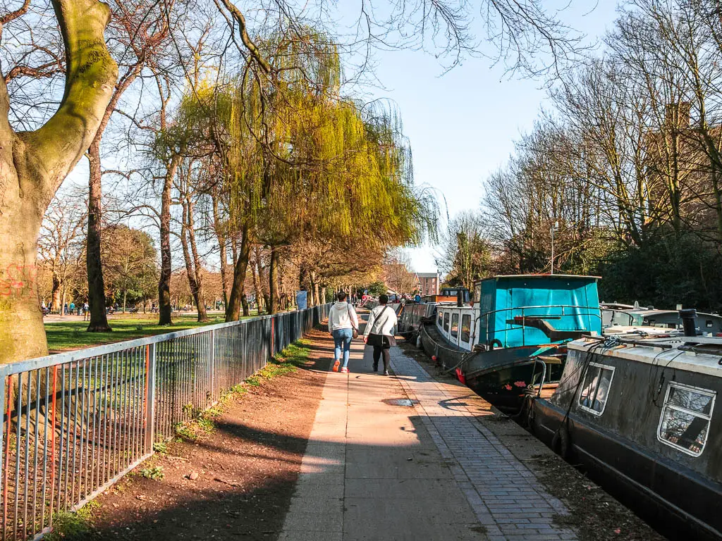 A long straight path with railings and park to the left and barges moored to the right. There are people walking on the path.