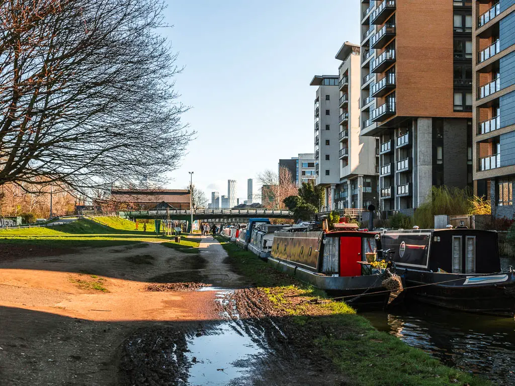 A path lined with grass on the walk along the Regents canal, with skyscrapers visible in the distance ahead. There are barges moored to the side, and apartment blocks on the other side of the canal.