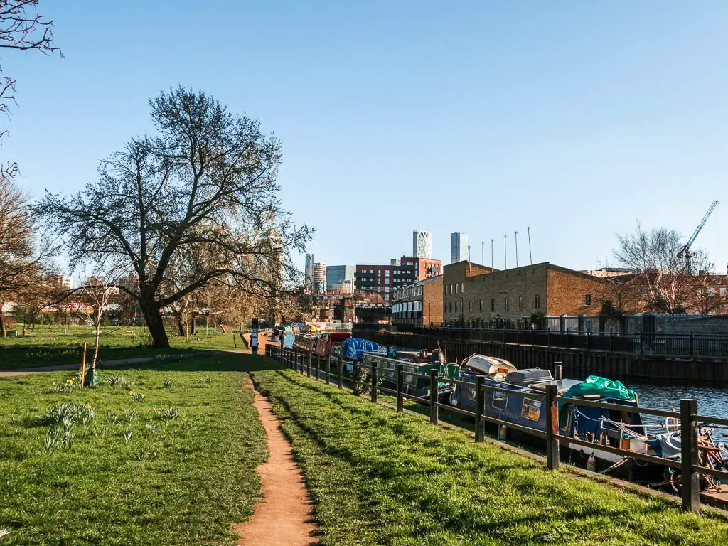 A carry path through the green, with black metal railings on the left, next to the canal. There are barges moored to the side. 