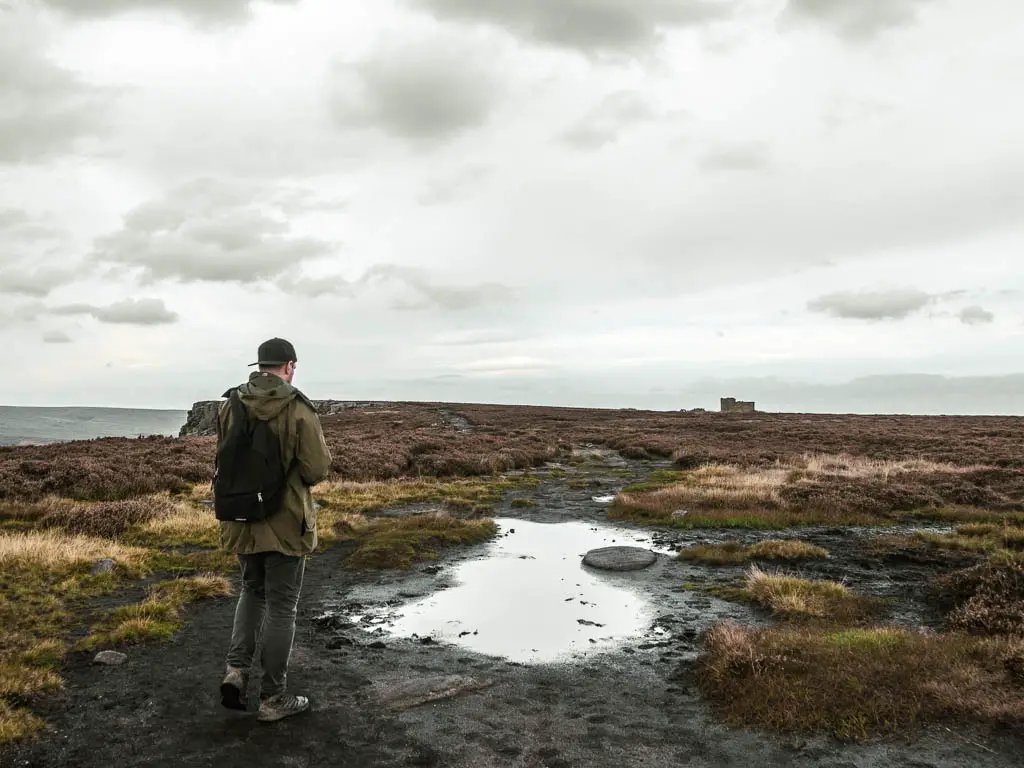 A man walking along a black coloured dirt path, with a big puddle ahead. The path is surround by grass and heather. There is a small building visible in the distance.
