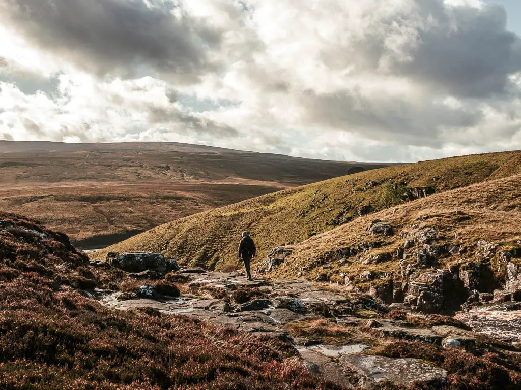 A man walking on some flat rocks in a dip in the hill, with a drop ahead, on the walk towards Cauldron snout.