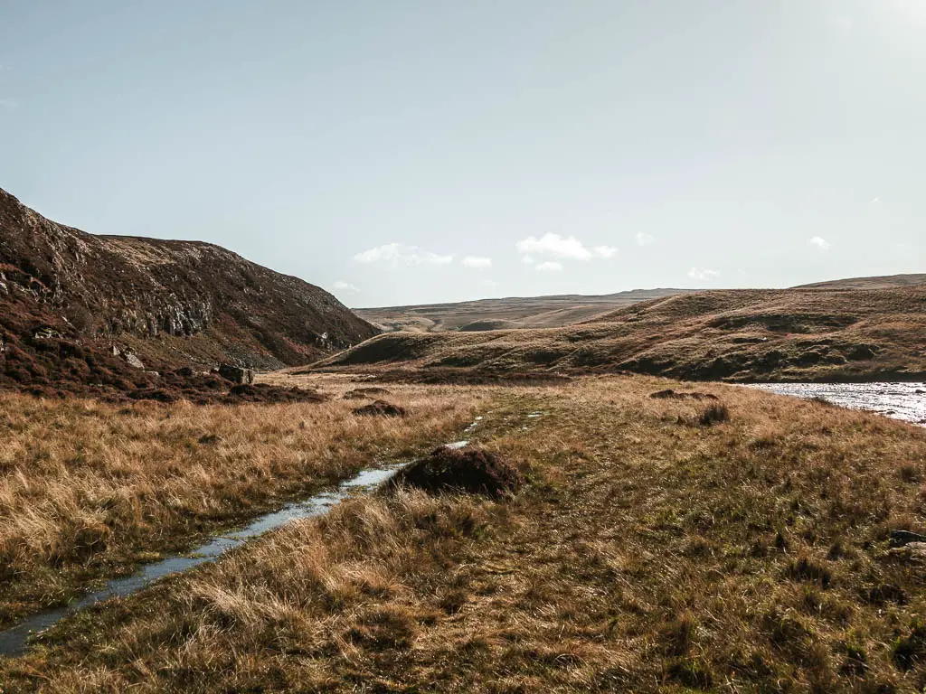 The trail covered in water, leading through the boggy ground, with hills ahead and to the left.