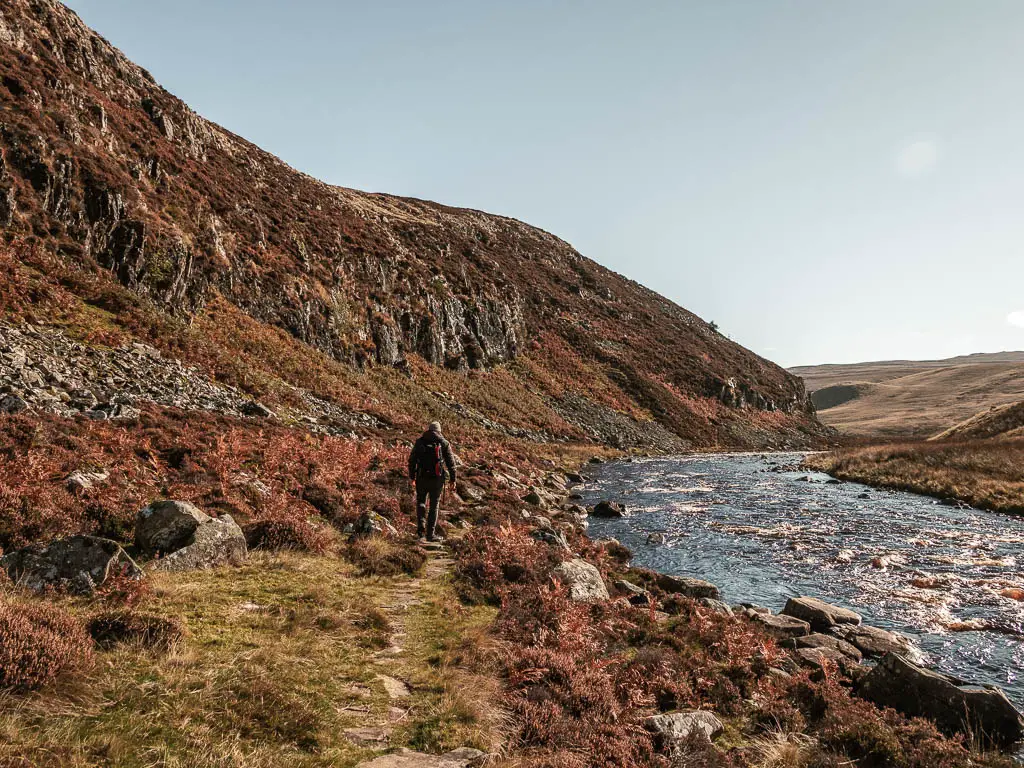 A man walking along the bottom of the rocky cliff face, with the river just to the right. The.ground is covered in rocks.