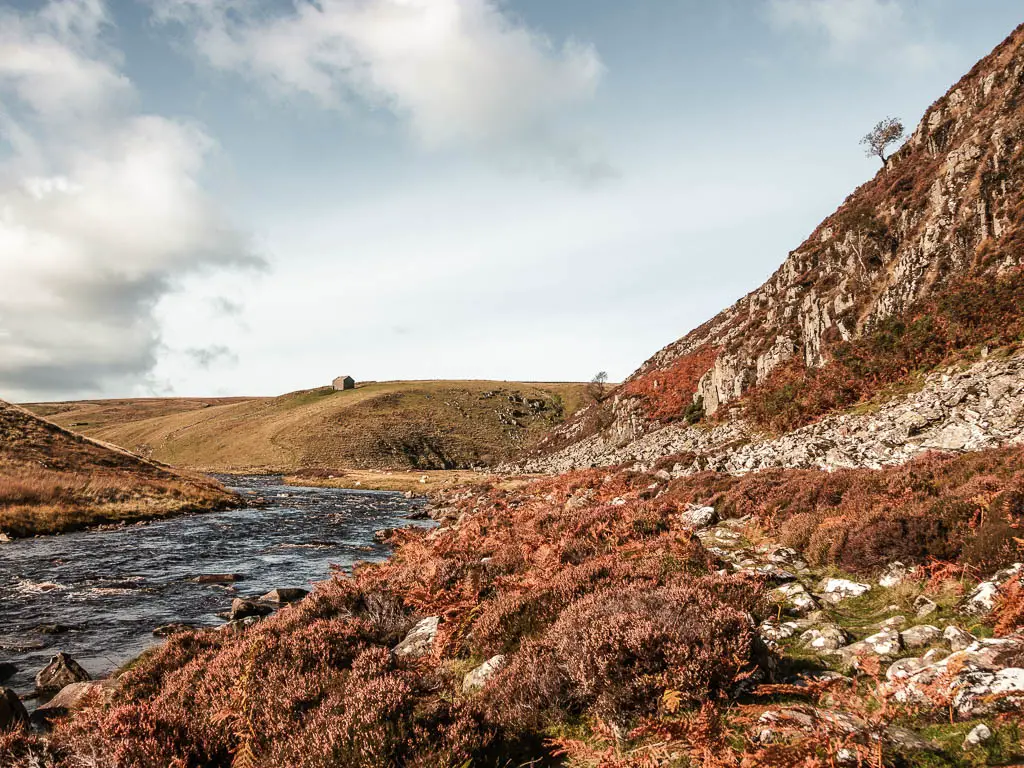 A river on the left, with a rocky cliff face to the right, and a rocky bush covered trail in between, on the circular Cauldron snout walk. There is a hill in the distance with a small hut on top.