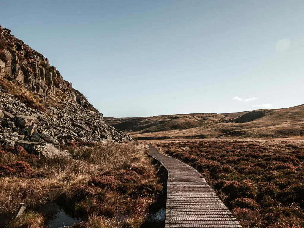 A wooden plan walkway across the boggy ground, with a rocky cliff face to the left.