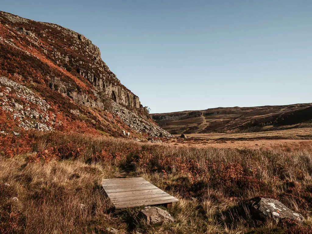 A small wooden plank walkway on the boggy ground, with a rocky cliff face to the left, and hills ahead to the right.