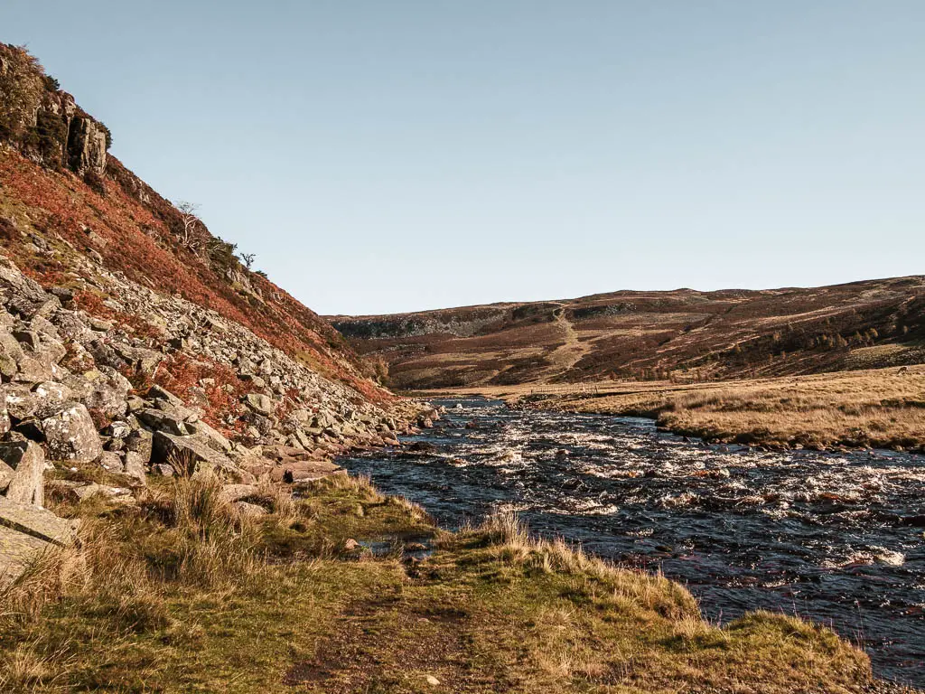 The deep blue almost black river flowing through the valley, with a hill covered in rocks to the left, on the Cauldron Snout circular walk.