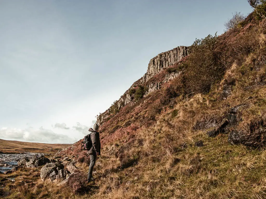 A man looking up to the rock cliff face along the Cauldron snout walk. The ground is covered in rugged overgrown grass and rocks. A bit of the river is visible to the left.