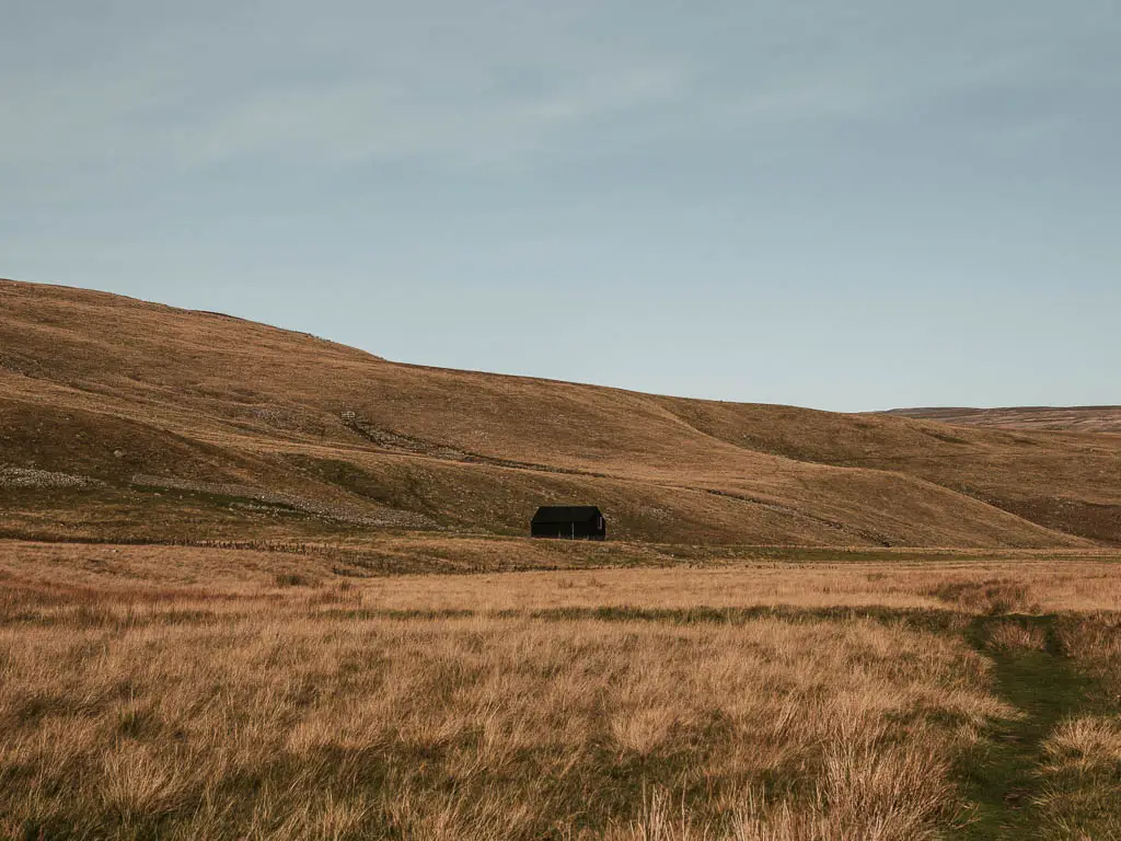 A small black hut on the other side of the field , with a hill rising up behind it, along the Cauldron Snout circular walk.