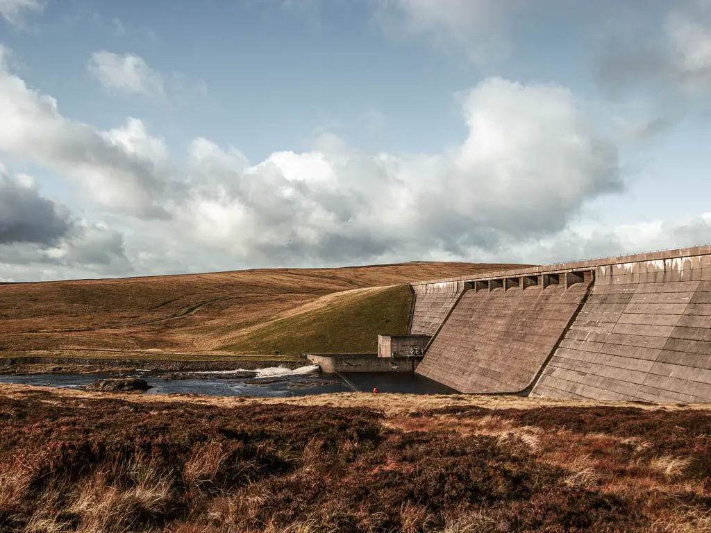 The cow green reservoir dam on the right, feeding into a small river the left. There is a hill on the other side of the water.