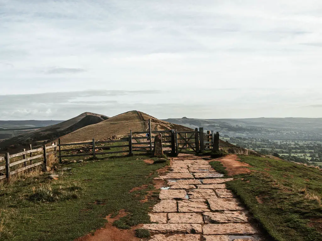 The stone paved path of the Great Ridge, leading to a wooden gate and fence, with hills in the distance.