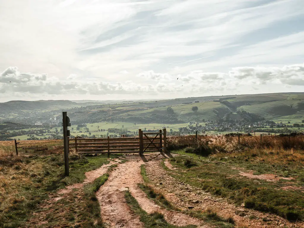 A dirt trail leading to a wooden fence and gate, with green fields and hills in the distance.