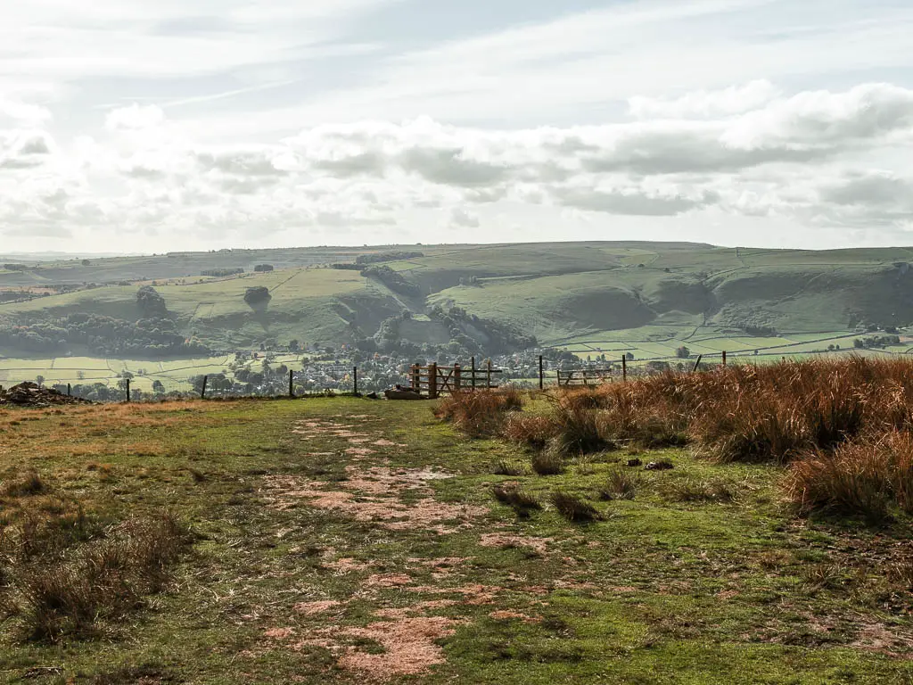A grass field with patches of dirt marking the trail, tifs of grass, and a fence on the other side. There are hills rising up in the distance.