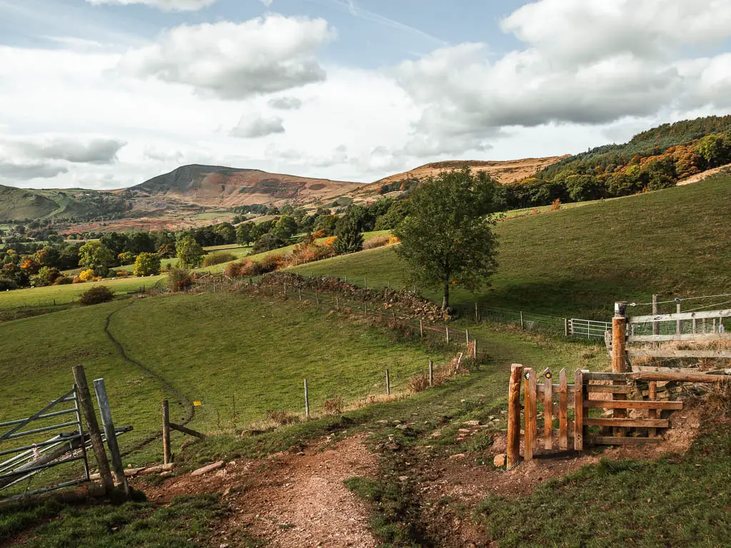 Walking down a dirt path, winding between grass hill fields, with a stone wall ahead, trees beyond that, then Mam Tor rising in the distance. 