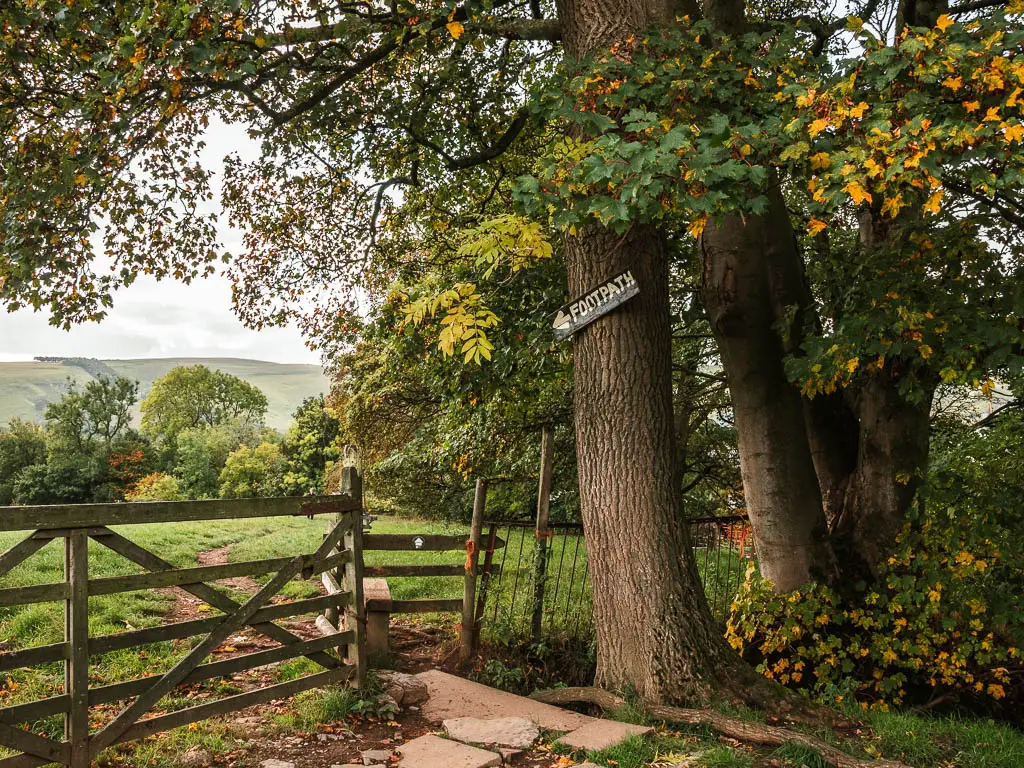 A sign saying 'footpath' Hugh on the tree trunk, pointing past a wooden fence and gate, into a field, on the circular walk route from Mam Tor.