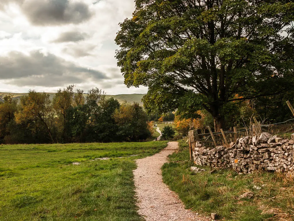 A gravel path with a grass field on the left and stone wall on the right. There are trees ahead on the other side of the field.