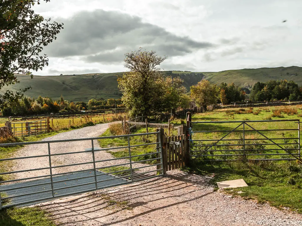 A wide gravel road, with a metal and wooden gate across it, and a grass field to the right.