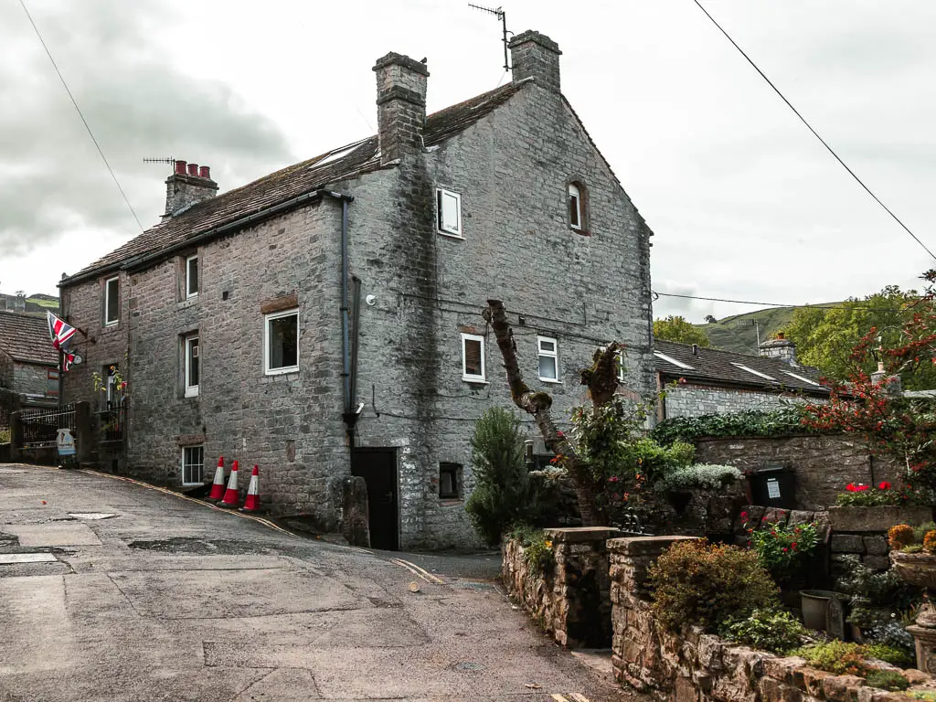 A large grey stone house in Castleton in the Peak District. There is a Union Jack flag hanging in front of it. To the right there is a flower front garden enclosed by a stone wall.