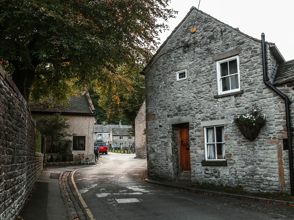 A road with a stone wall on the left and stone house with a brown door on the right in Castleton.