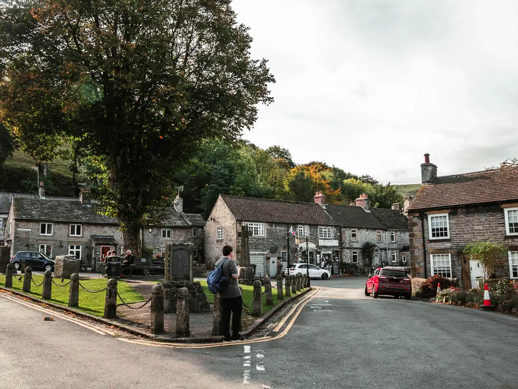 The road splitting around a green town square, with stone houses on the other side and a Brock house on the right. There is a person walking along the side of the square.