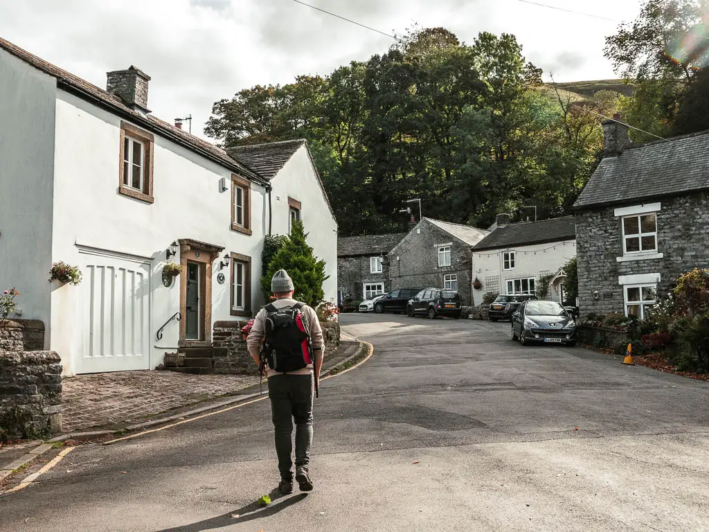A man walking up a residential road. There is a white coloured house on the left, and grey and white stone houses to the right.