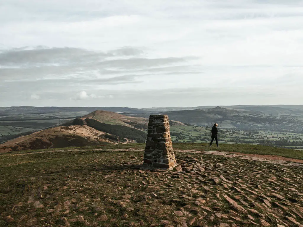 A stoney ground with a trig point, on the top of Mam Tor and a person walking along the other side. The Great Ridge is in the distance on the left.