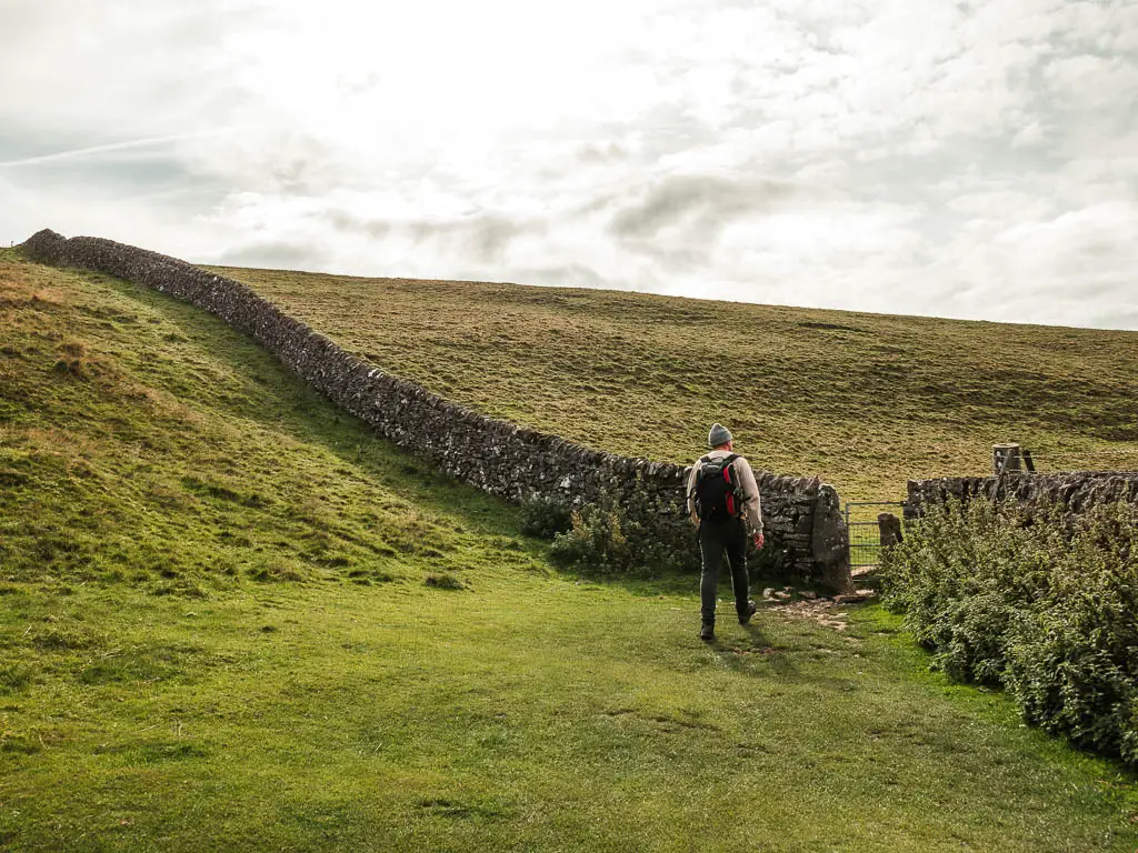 A stone wall running down the green grass hill, with a small gate in the middle of it. There is a man walking towards the gate.