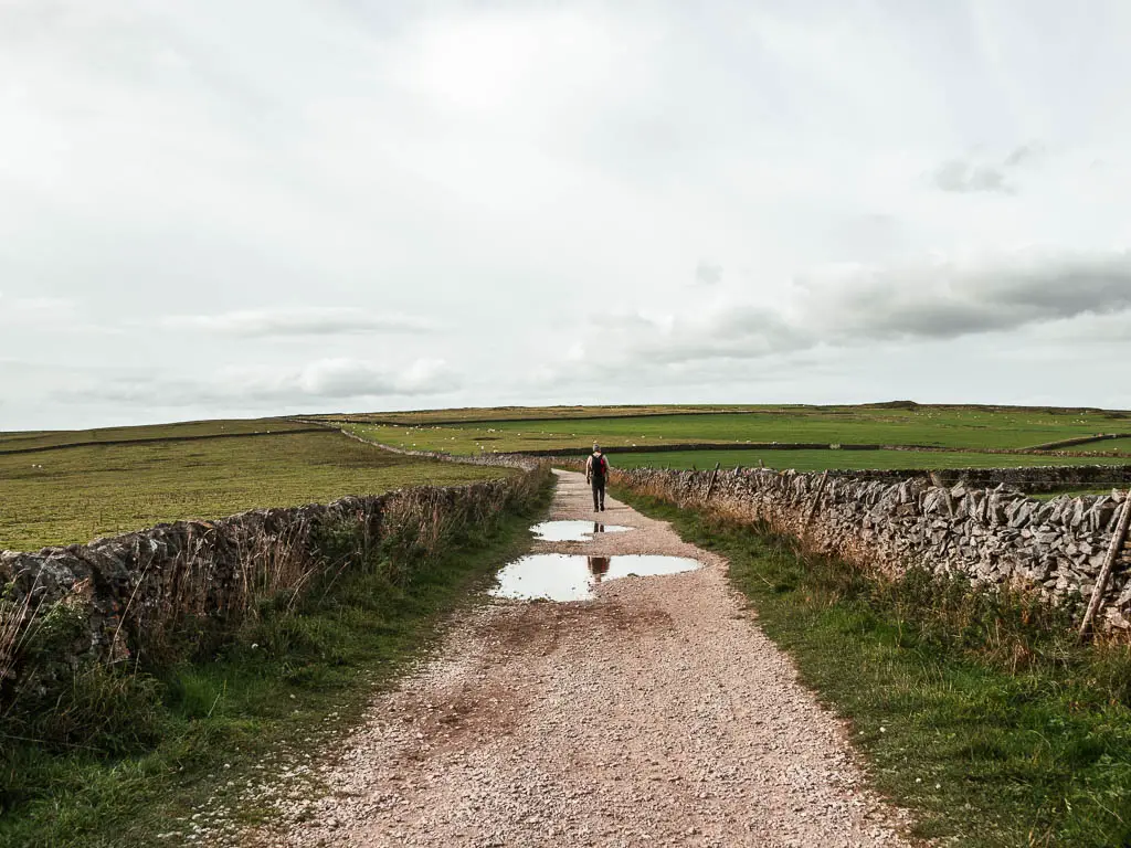 A long wide gravel path, lines with a a stone wall on either side. There are puddles in the path and man walking along it in the distance.
