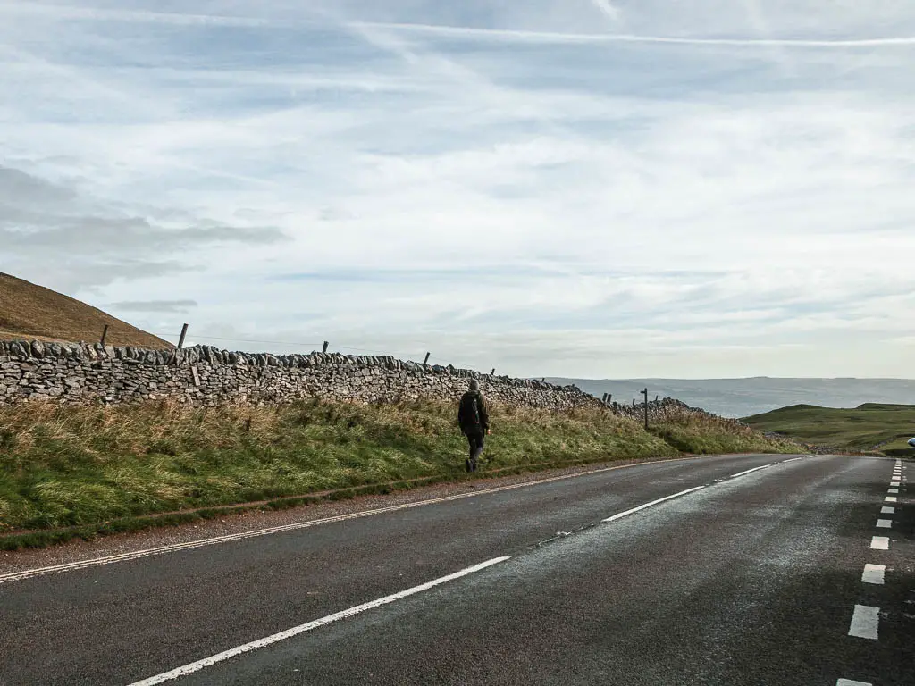 A man walking along the side of a road, next to a stone wall.