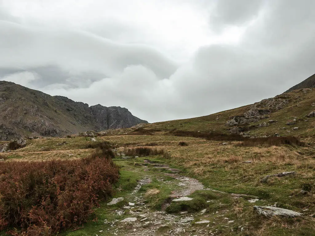 A grass path of ground with some rocks and a hill ahead to the left.