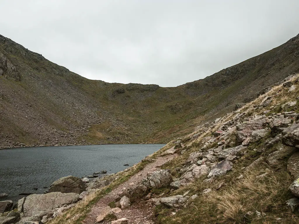 A narrow path leading around the right side of Goats water which is surround by a big steep hill.