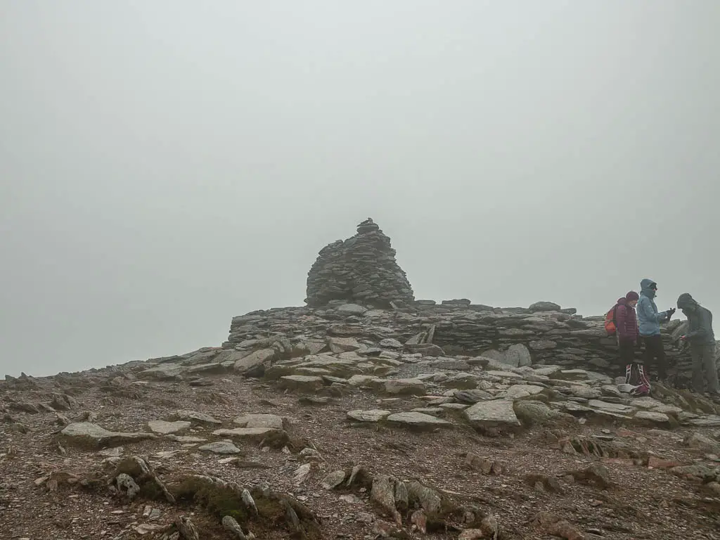 The rocky peak of the Old Man of Coniston, with a few people walking to the right. The is mist and fog all around.