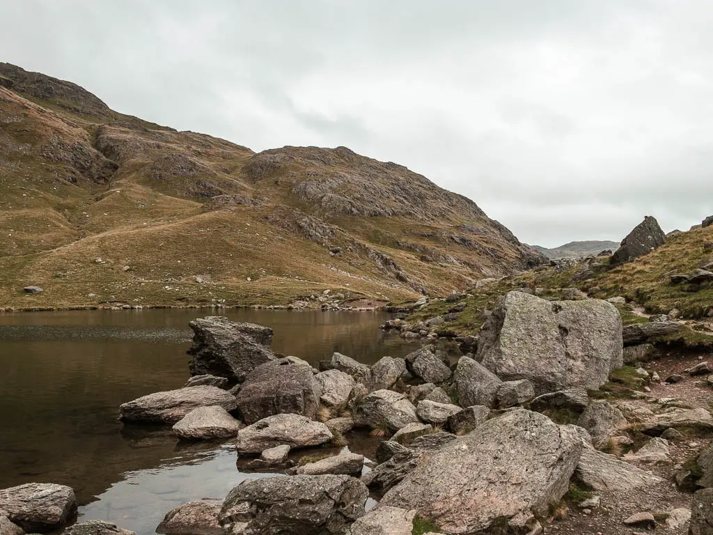 Large rocks lining the right side of a small lake, with a big hill on the other side.