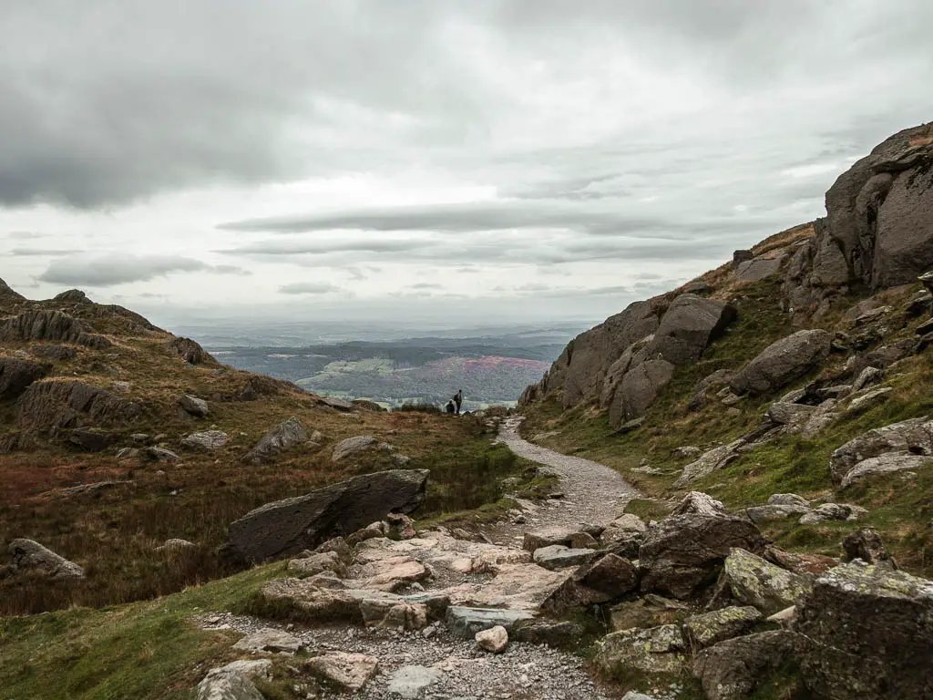 A path winding ahead, without a gap in the hill and rocks ahead, and a view  to the fields on the valley in the distance.