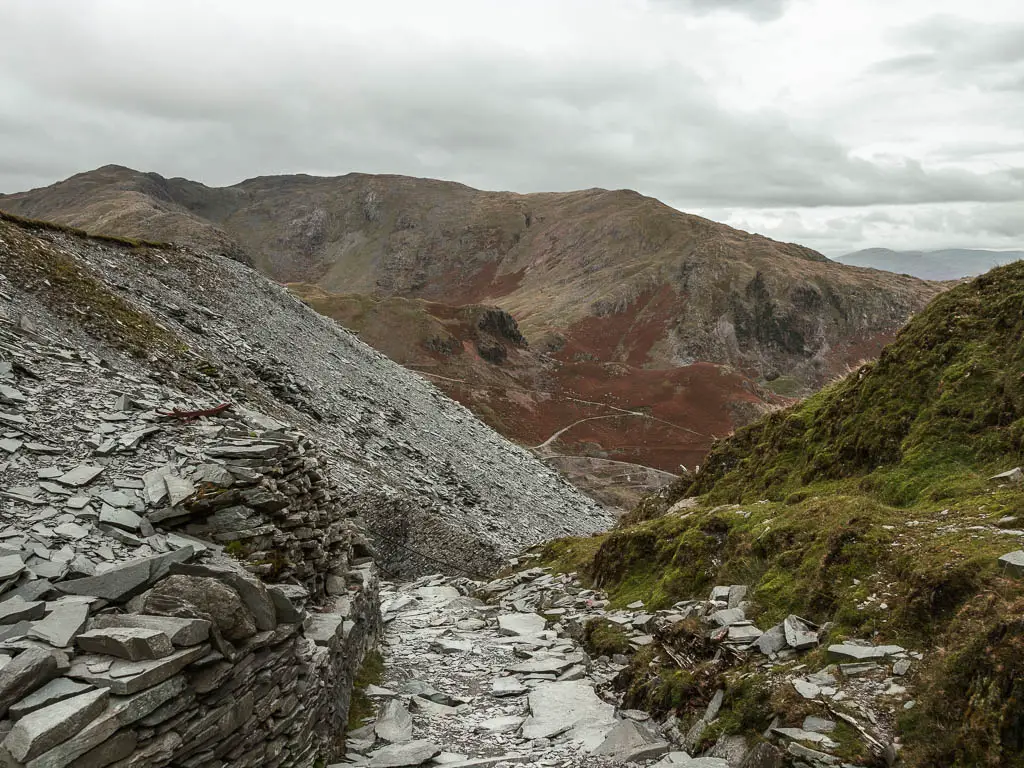 A path of slate rock with a grass hill on the right and crumbling slate rock wall on the left.