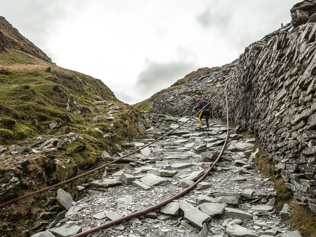 A man walking under two thick cables running across the slate rock trai,l on the Old Man of Coniston circular walk.