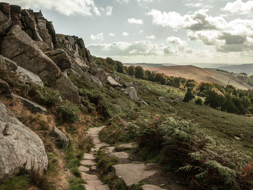 A stone paved trail on the walk below Stanage Edge. The rock face wall is to the left, with some woodland trees ahead.